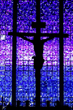 A silhouette of a crucifix is set against a backdrop of a stained glass window featuring a mosaic of deep blues and purples.
