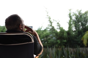 A person is seated on a chair outdoors, appearing to be deep in thought. The background features lush green trees and plants, suggesting a serene garden or park setting. The lighting is soft, contributing to a peaceful atmosphere.