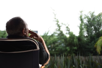 A person is seated on a chair outdoors, appearing to be deep in thought. The background features lush green trees and plants, suggesting a serene garden or park setting. The lighting is soft, contributing to a peaceful atmosphere.