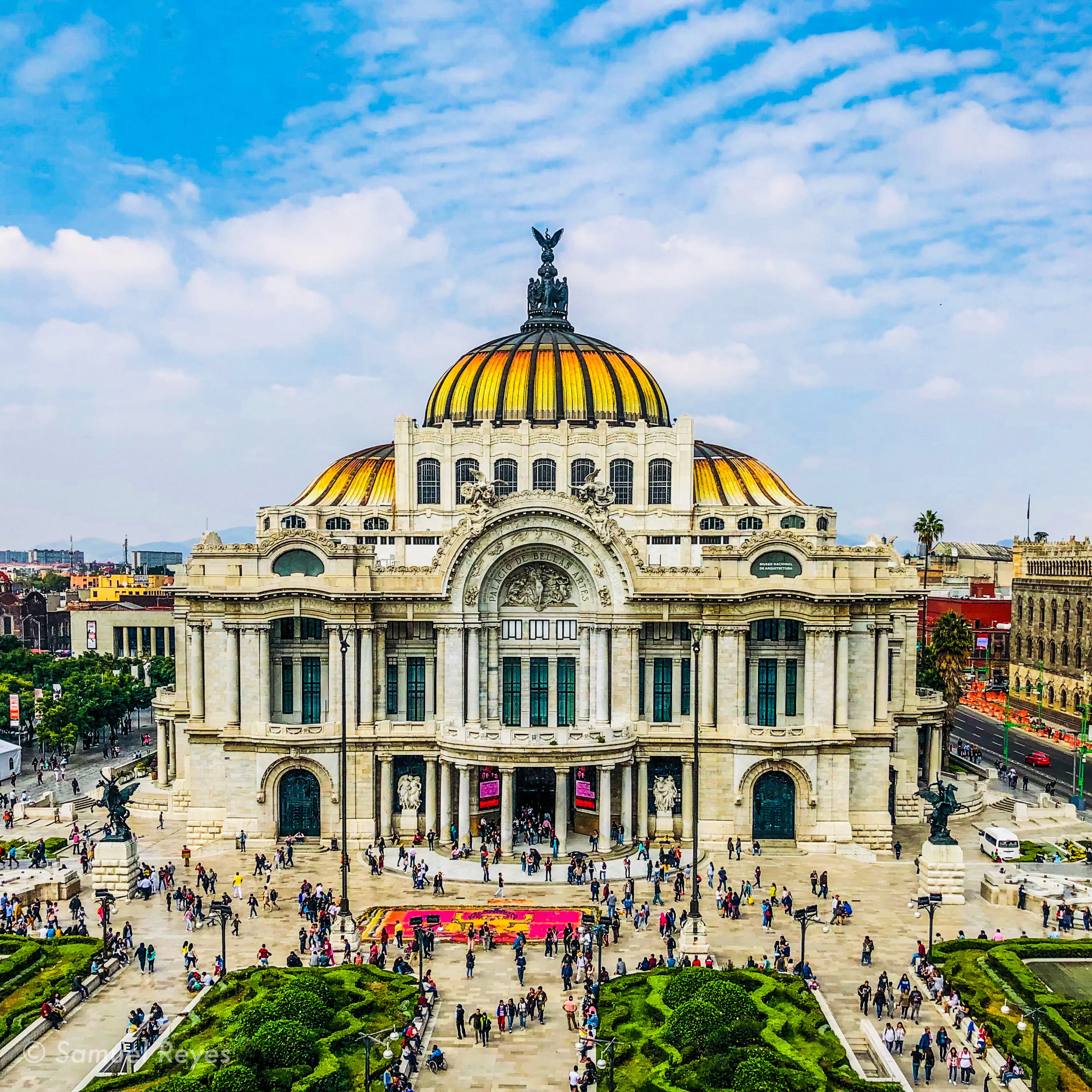 The Palacio de Bellas Artes in Mexico City seen from above, its cream marble facade crowned by a yellow-and-orange tiled dome, with crowds on the plaza below