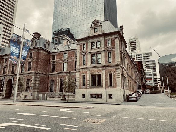 A historic red brick building with classic architectural features is situated in an urban area. Modern skyscrapers rise in the background, creating a contrast between old and new architecture. Banners are visible on lamp posts, and several parked cars line the street beside the building.