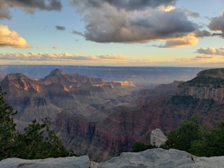 A vast landscape of the Grand Canyon with towering rock formations illuminated by soft sunlight. The rugged terrain is complemented by lush greenery at the foreground and dramatic cloud formations in the sky.
