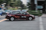 A maroon taxi with colorful pixel-like patterns on the side is moving at speed through an urban area. The background shows a motion blur effect with hints of greenery and buildings, suggesting it's in a city.