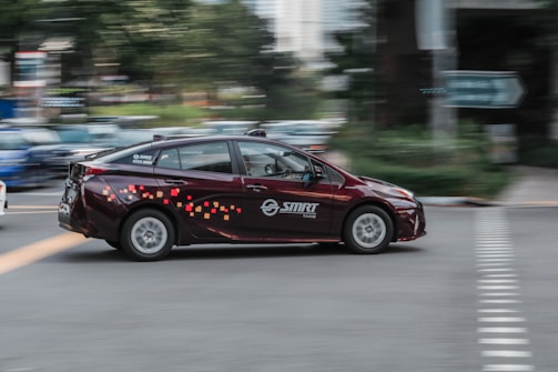 A maroon taxi with colorful pixel-like patterns on the side is moving at speed through an urban area. The background shows a motion blur effect with hints of greenery and buildings, suggesting it's in a city.