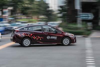 A maroon taxi with colorful pixel-like patterns on the side is moving at speed through an urban area. The background shows a motion blur effect with hints of greenery and buildings, suggesting it's in a city.