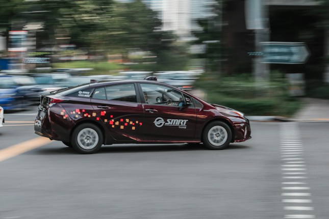 A maroon taxi with colorful pixel-like patterns on the side is moving at speed through an urban area. The background shows a motion blur effect with hints of greenery and buildings, suggesting it's in a city.