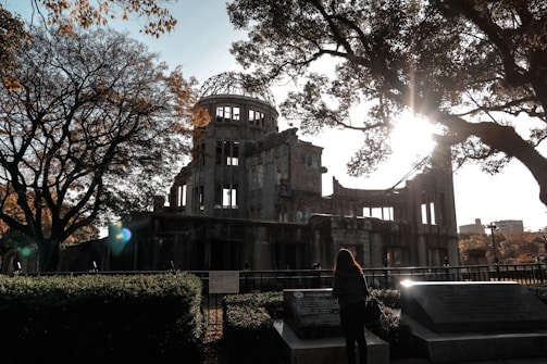 A person stands in front of a historic, partially destroyed building surrounded by trees. The sun is shining brightly through the branches of the trees, casting a mixture of light and shadow on the scene. There are informational plaques or monuments in front of the person, likely describing the site's significance.