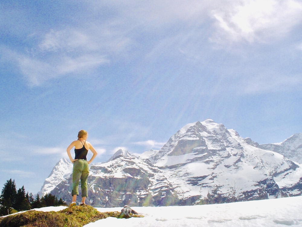 woman with both arms akimbo standing on grass rock on snow field near glacier mountains