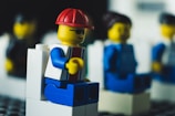 A close-up view of a Lego figure wearing a red cap and sunglasses, seated on a white block. Surrounding figures in the background are out of focus, suggesting a social or group setting.