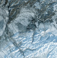 A satellite view of a rugged, mountainous landscape with intricate patterns of ridges and valleys. The terrain is covered in snow, creating a light blue and white palette. There are visible rivers and streams cutting through the land, adding contrast to the snowy expanses.