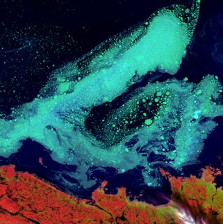 A mesmerizing satellite image captures a dramatic view of Earth's natural formations. Bright turquoise seas act as a striking contrast against deep blue waters, showing swirling patterns that resemble ocean currents or algae blooms. In the lower part, rugged red and orange landmasses highlight the coastline, offering a vivid display of geographical features.