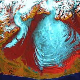 A vivid satellite image highlighting a large glacial landscape with striking swirls of ice. The ice appears in vibrant blues and whites, contrasted starkly by surrounding red and brown rocky terrains. Snow-capped mountains frame the upper portion of the image, while the bottom edge reveals a dark blue body of water along the coastline. The flowing patterns suggest a dynamic movement of ice across the terrain.