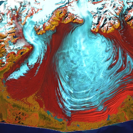 A vivid satellite image highlighting a large glacial landscape with striking swirls of ice. The ice appears in vibrant blues and whites, contrasted starkly by surrounding red and brown rocky terrains. Snow-capped mountains frame the upper portion of the image, while the bottom edge reveals a dark blue body of water along the coastline. The flowing patterns suggest a dynamic movement of ice across the terrain.