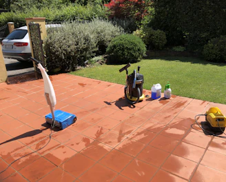 A sunlit patio with terracotta tiles is adorned with cleaning equipment, including a wet and dry vacuum, containers of cleaning solutions, and a pressure washer. A portion of a white car is visible near a hedge-lined driveway, while lush green vegetation and a manicured lawn provide a backdrop.