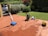 A sunlit patio with terracotta tiles is adorned with cleaning equipment, including a wet and dry vacuum, containers of cleaning solutions, and a pressure washer. A portion of a white car is visible near a hedge-lined driveway, while lush green vegetation and a manicured lawn provide a backdrop.
