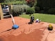 A sunlit patio with terracotta tiles is adorned with cleaning equipment, including a wet and dry vacuum, containers of cleaning solutions, and a pressure washer. A portion of a white car is visible near a hedge-lined driveway, while lush green vegetation and a manicured lawn provide a backdrop.