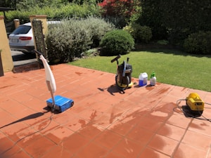 A sunlit patio with terracotta tiles is adorned with cleaning equipment, including a wet and dry vacuum, containers of cleaning solutions, and a pressure washer. A portion of a white car is visible near a hedge-lined driveway, while lush green vegetation and a manicured lawn provide a backdrop.