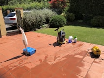 A sunlit patio with terracotta tiles is adorned with cleaning equipment, including a wet and dry vacuum, containers of cleaning solutions, and a pressure washer. A portion of a white car is visible near a hedge-lined driveway, while lush green vegetation and a manicured lawn provide a backdrop.