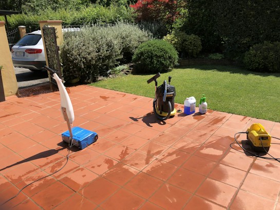A sunlit patio with terracotta tiles is adorned with cleaning equipment, including a wet and dry vacuum, containers of cleaning solutions, and a pressure washer. A portion of a white car is visible near a hedge-lined driveway, while lush green vegetation and a manicured lawn provide a backdrop.