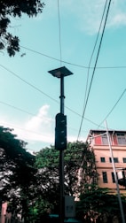 A tall streetlight pole accompanied by a solar panel, surrounded by trees and a few buildings in the background. Several power lines crisscross the scene under a bright sky. The foliage appears lush and green, contrasting with the urban elements.
