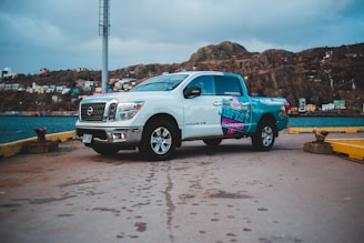 A white pickup truck with colorful decals is parked on a concrete dock near the water. In the background, there is a rocky hill with several colorful houses scattered across it under a cloudy sky. The truck is positioned sideways, showcasing its side and front.