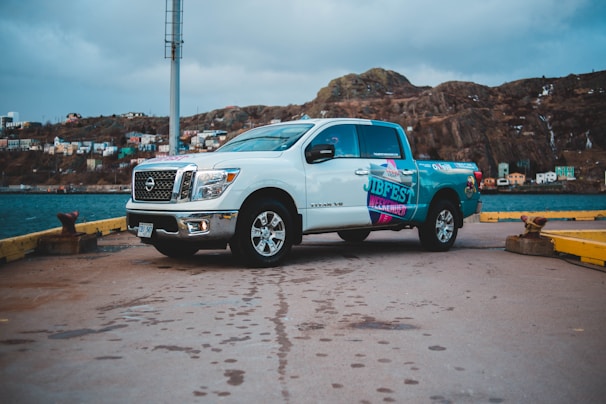 A white pickup truck with colorful decals is parked on a concrete dock near the water. In the background, there is a rocky hill with several colorful houses scattered across it under a cloudy sky. The truck is positioned sideways, showcasing its side and front.
