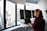 A person wearing headphones is working at a standing desk with multiple computer monitors displaying code. The room features large windows with a view of city buildings outside. The person is wearing a red plaid shirt and appears focused on their work.