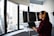 A person wearing headphones is working at a standing desk with multiple computer monitors displaying code. The room features large windows with a view of city buildings outside. The person is wearing a red plaid shirt and appears focused on their work.