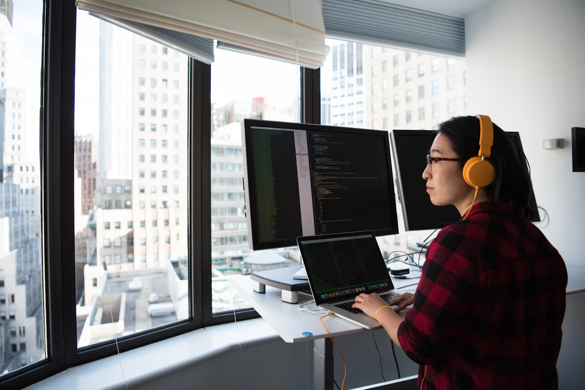 An engineer in orange headphones works at a desk with code on two monitors and a laptop beside high-rise windows
