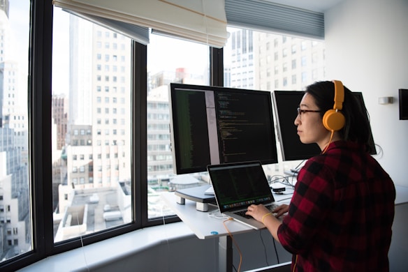A person wearing headphones is working at a standing desk with multiple computer monitors displaying code. The room features large windows with a view of city buildings outside. The person is wearing a red plaid shirt and appears focused on their work.