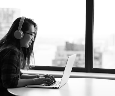 woman wearing headphones using laptop beside the glass window grayscale photo