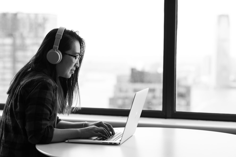 woman wearing headphones using laptop beside the glass window grayscale photo