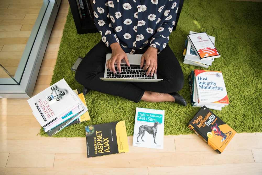 woman using laptop sitting on carpet surrounded with books