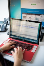 A person is typing on a laptop with a red cover. The laptop screen displays an audio editing software with visible waveforms. In the background, there is a larger computer monitor showing a website with various tab previews. A black and gray desk phone is also present on the left.