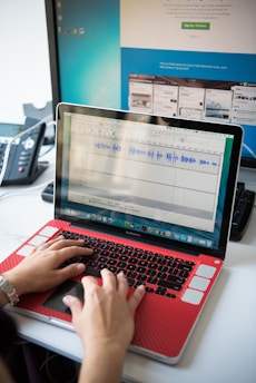 A person is typing on a laptop with a red cover. The laptop screen displays an audio editing software with visible waveforms. In the background, there is a larger computer monitor showing a website with various tab previews. A black and gray desk phone is also present on the left.