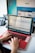 A person is typing on a laptop with a red cover. The laptop screen displays an audio editing software with visible waveforms. In the background, there is a larger computer monitor showing a website with various tab previews. A black and gray desk phone is also present on the left.