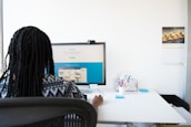 A person with braided hair is sitting at a desk working on a computer monitor displaying a website. The desk is clean and organized, featuring a pen holder with stationery, a chair with a mesh back, and a wall calendar displaying an image of food.