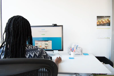A person with braided hair is sitting at a desk working on a computer monitor displaying a website. The desk is clean and organized, featuring a pen holder with stationery, a chair with a mesh back, and a wall calendar displaying an image of food.