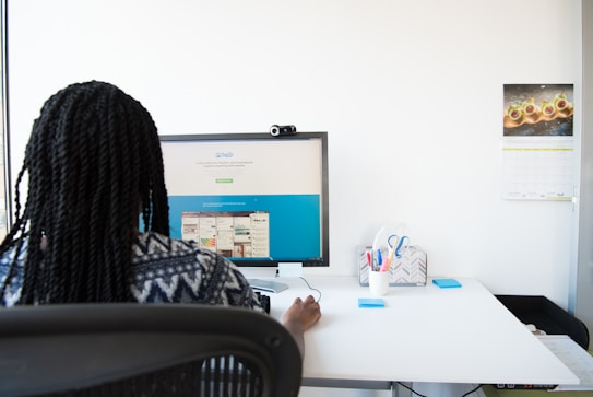 A person with braided hair is sitting at a desk working on a computer monitor displaying a website. The desk is clean and organized, featuring a pen holder with stationery, a chair with a mesh back, and a wall calendar displaying an image of food.