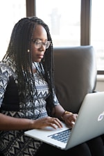 A person with braided hair is sitting on a sofa, using a laptop. They appear focused on their work in a well-lit room with large windows in the background.