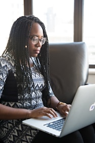 A person with braided hair is sitting on a sofa, using a laptop. They appear focused on their work in a well-lit room with large windows in the background.