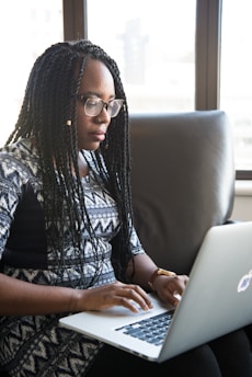 A person with braided hair is sitting on a sofa, using a laptop. They appear focused on their work in a well-lit room with large windows in the background.