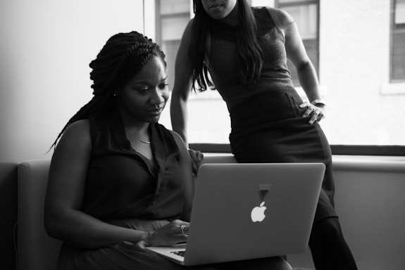 Two women are in an office setting looking at a laptop. One is seated and focused on the screen while the other stands beside her, leaning slightly over, with one hand on her hip. Natural light from a window illuminates the room, giving the photo a professional atmosphere.