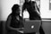 Two women are in an office setting looking at a laptop. One is seated and focused on the screen while the other stands beside her, leaning slightly over, with one hand on her hip. Natural light from a window illuminates the room, giving the photo a professional atmosphere.
