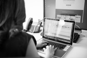 A person is seated at a desk working on a laptop. The laptop screen displays audio editing software with visible waveforms. A large monitor in the background shows a website with various sections. There is a telephone next to the monitor, and papers or notes are scattered on the desk.