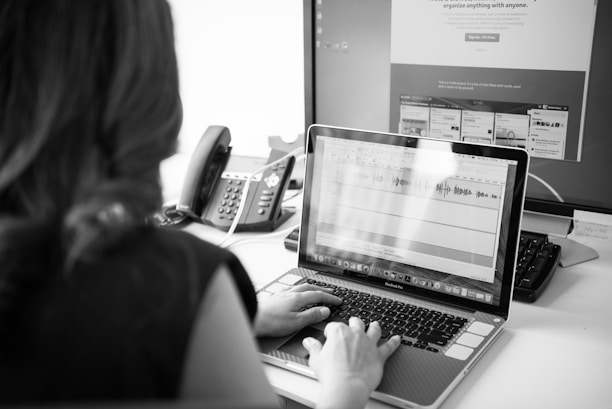 A person is seated at a desk working on a laptop. The laptop screen displays audio editing software with visible waveforms. A large monitor in the background shows a website with various sections. There is a telephone next to the monitor, and papers or notes are scattered on the desk.