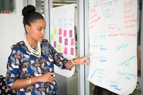 A woman with her hair tied up and wearing a patterned blouse is standing next to a whiteboard. She is pointing at a flowchart on the board while holding a marker. There are colorful sticky notes in the background, indicating a brainstorming or planning session.