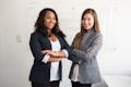 Two women are standing confidently with their arms crossed in a professional setting. They are dressed in business attire, consisting of blazers and light-colored tops. The background includes a whiteboard with some faint text written on it.