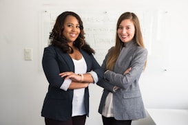 Two women are standing confidently with their arms crossed in a professional setting. They are dressed in business attire, consisting of blazers and light-colored tops. The background includes a whiteboard with some faint text written on it.