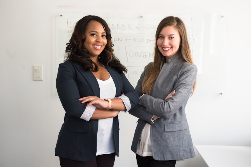 Two women are standing confidently with their arms crossed in a professional setting. They are dressed in business attire, consisting of blazers and light-colored tops. The background includes a whiteboard with some faint text written on it.